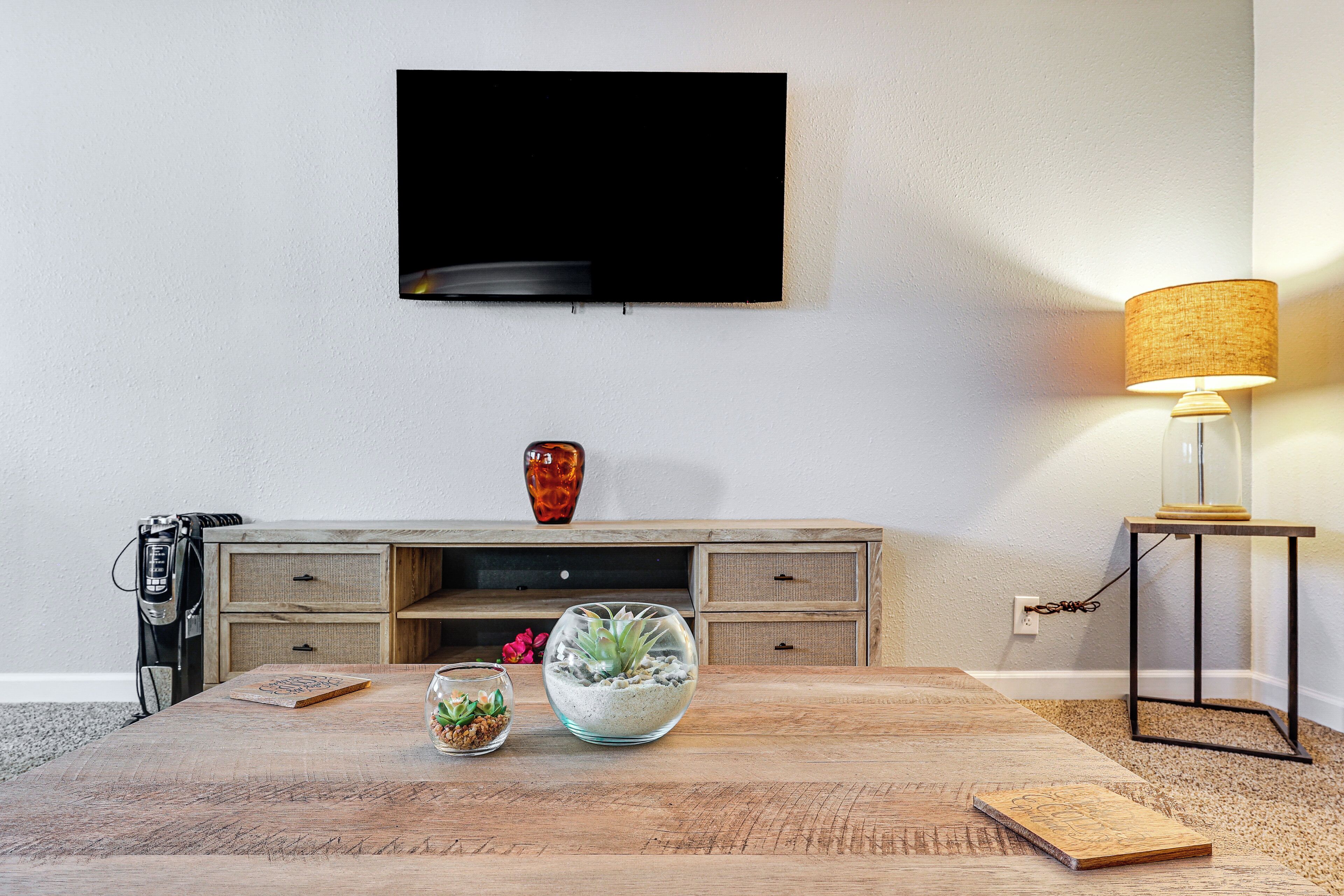 Una sala de estar con una mesa de centro de madera, un televisor de pantalla plana montado en la pared, una lámpara y una pequeña planta.