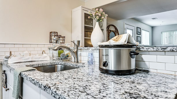 Una cocina con encimera de granito, una olla de acero inoxidable, un lavamanos y un armario blanco.