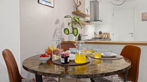 Uma sala de jantar com uma mesa redonda, cadeiras, uma planta e uma cozinha ao fundo.