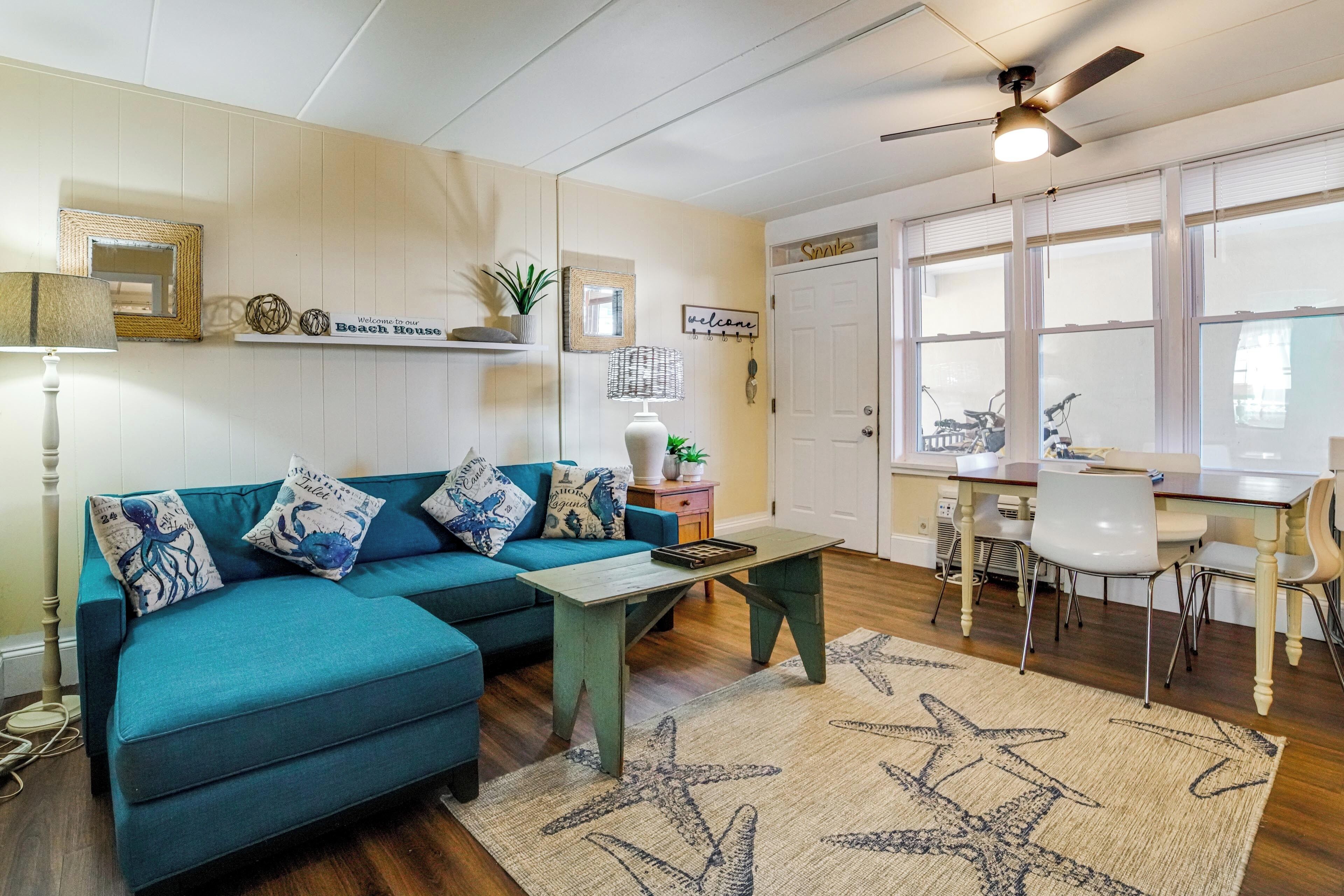 Un sofá azul verdoso con almohadones de estrella de mar, una mesa de centro de madera, un tapete con motivos de estrella de mar, un ventilador de techo y un cartel que dice 'Welcome to our Beach House'.