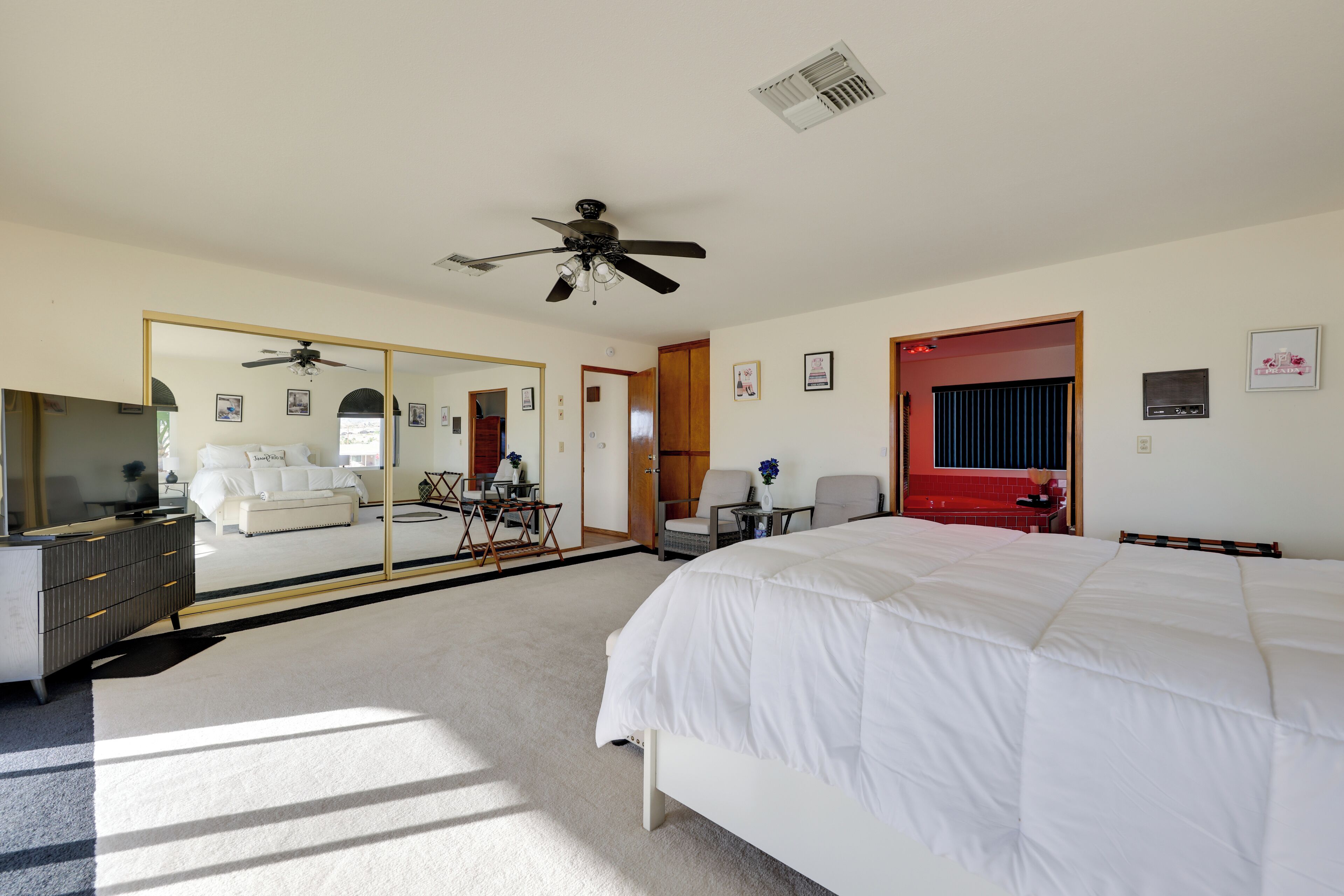 A bedroom with a large bed, a ceiling fan, a mirror, a dresser, and a red door.