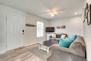 Un salon avec une porte blanche, un ventilateur de plafond, un téléviseur à écran plat, un canapé avec des coussins turquoise et bruns, et un tapis à motifs.