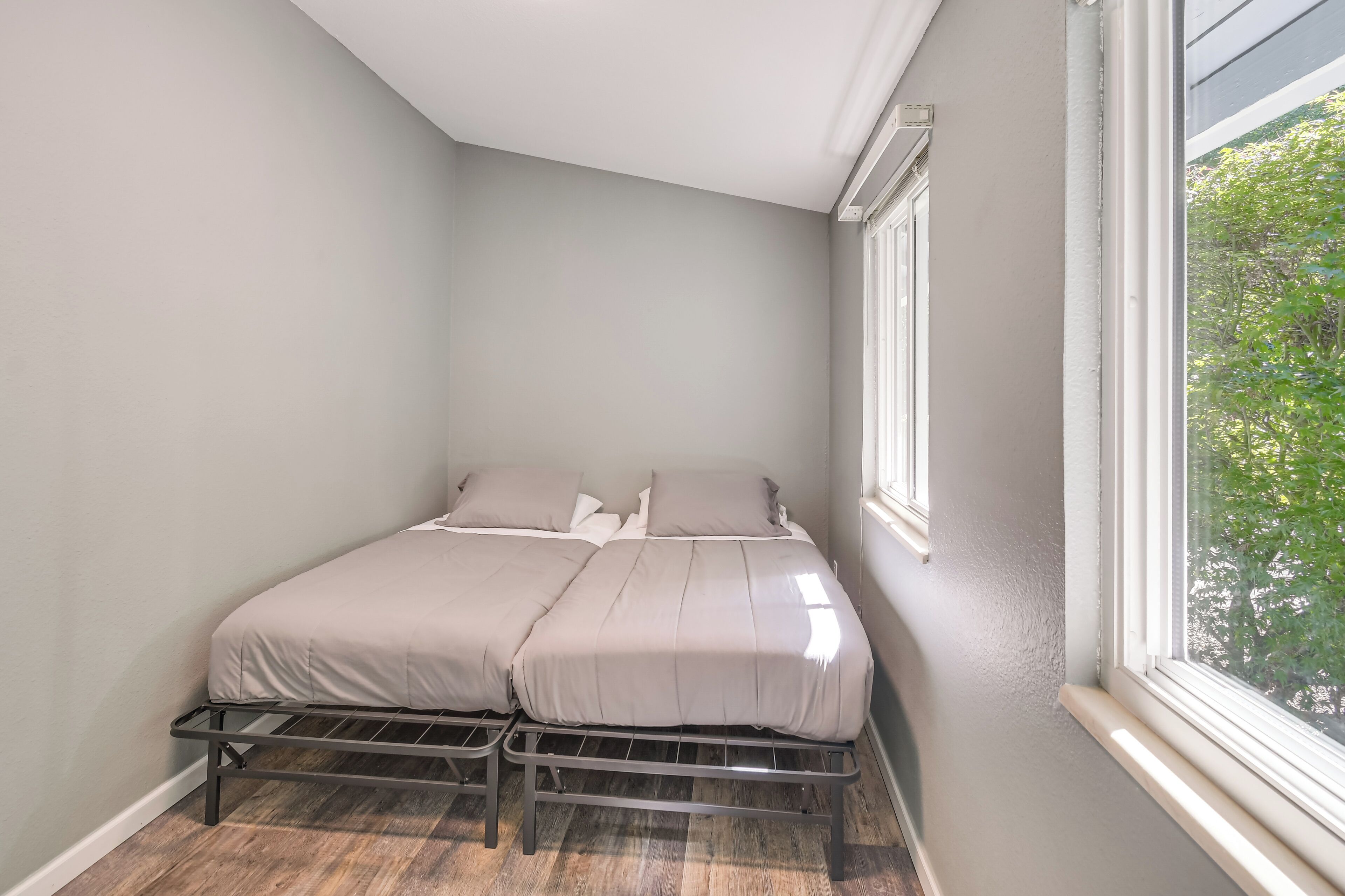 A small, single-bed room with a metal bed frame, a window with a view of greenery, and a wooden floor.