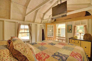 A bedroom with a quilted bedspread, a television mounted on the ceiling, a dresser, and a window with curtains.