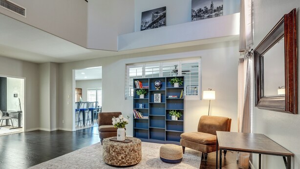 A spacious living area with a dark wood floor, a round coffee table with a vase, a bookshelf, and a large mirror. - Modern Family Oasis Pool Game Room (Maricopa)