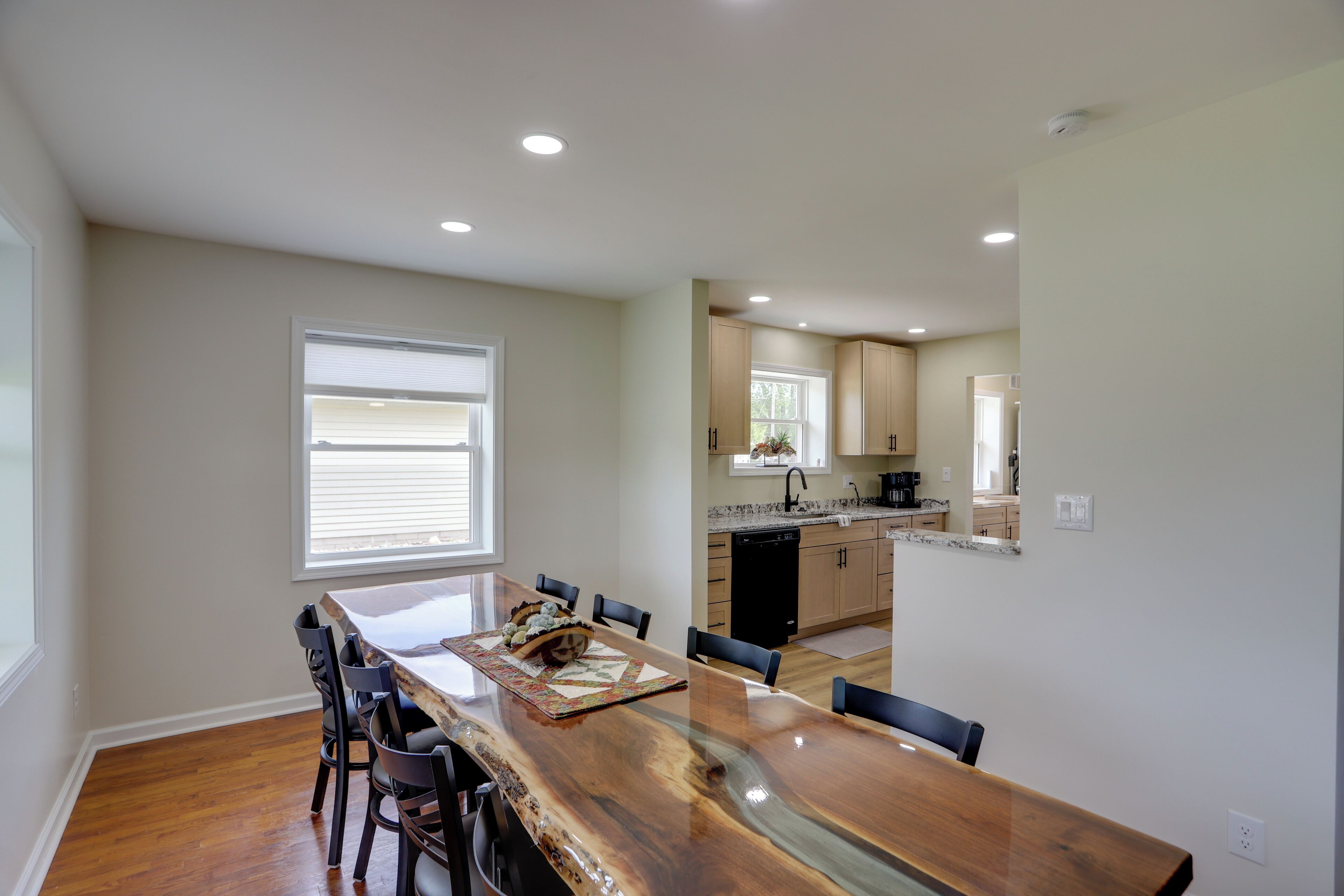 A dining area with a long wooden table, chairs, a window, and a kitchen in the background.