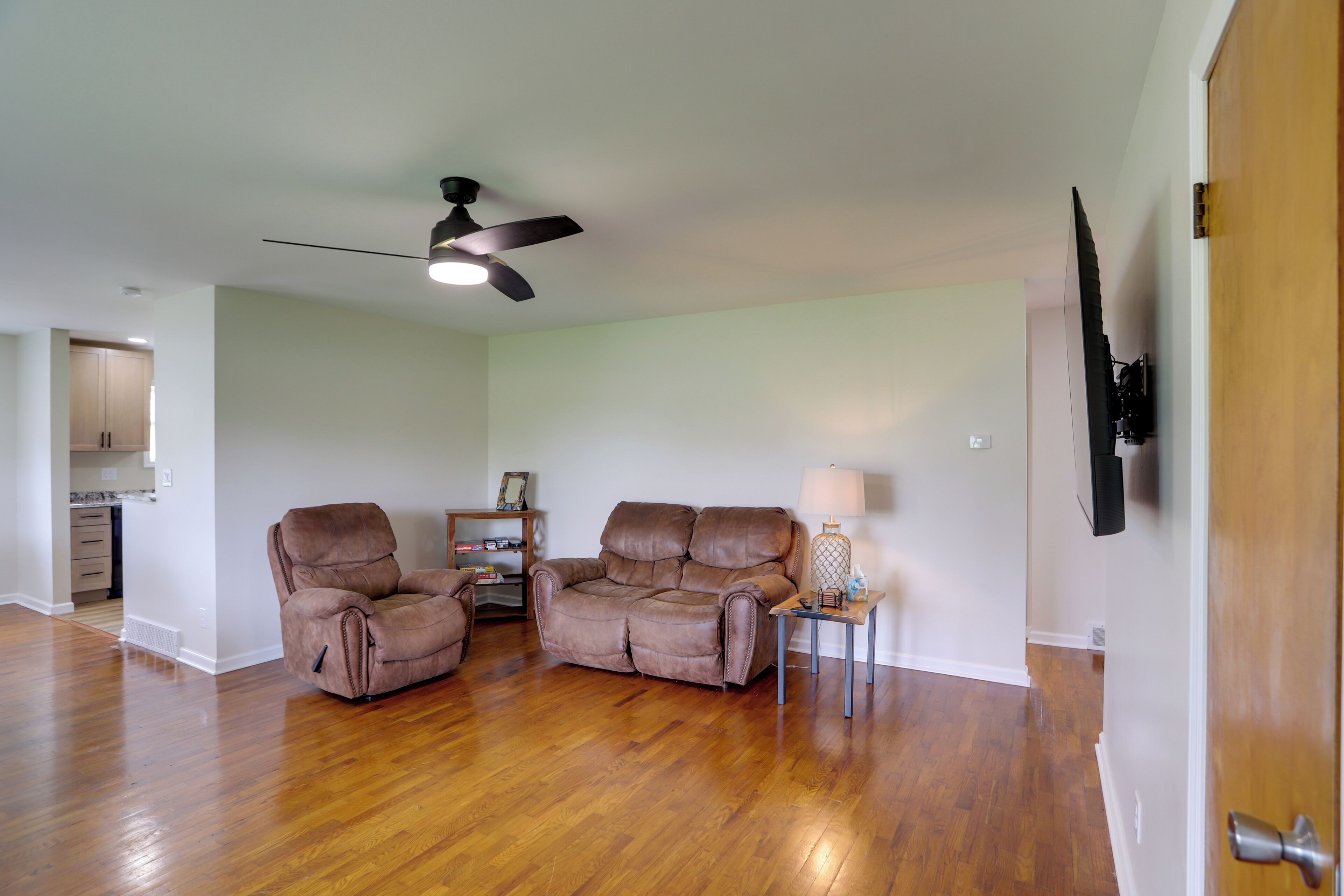 A spacious living room with hardwood flooring, two leather recliners, a ceiling fan, a side table with a lamp, and a television mounted on the wall.