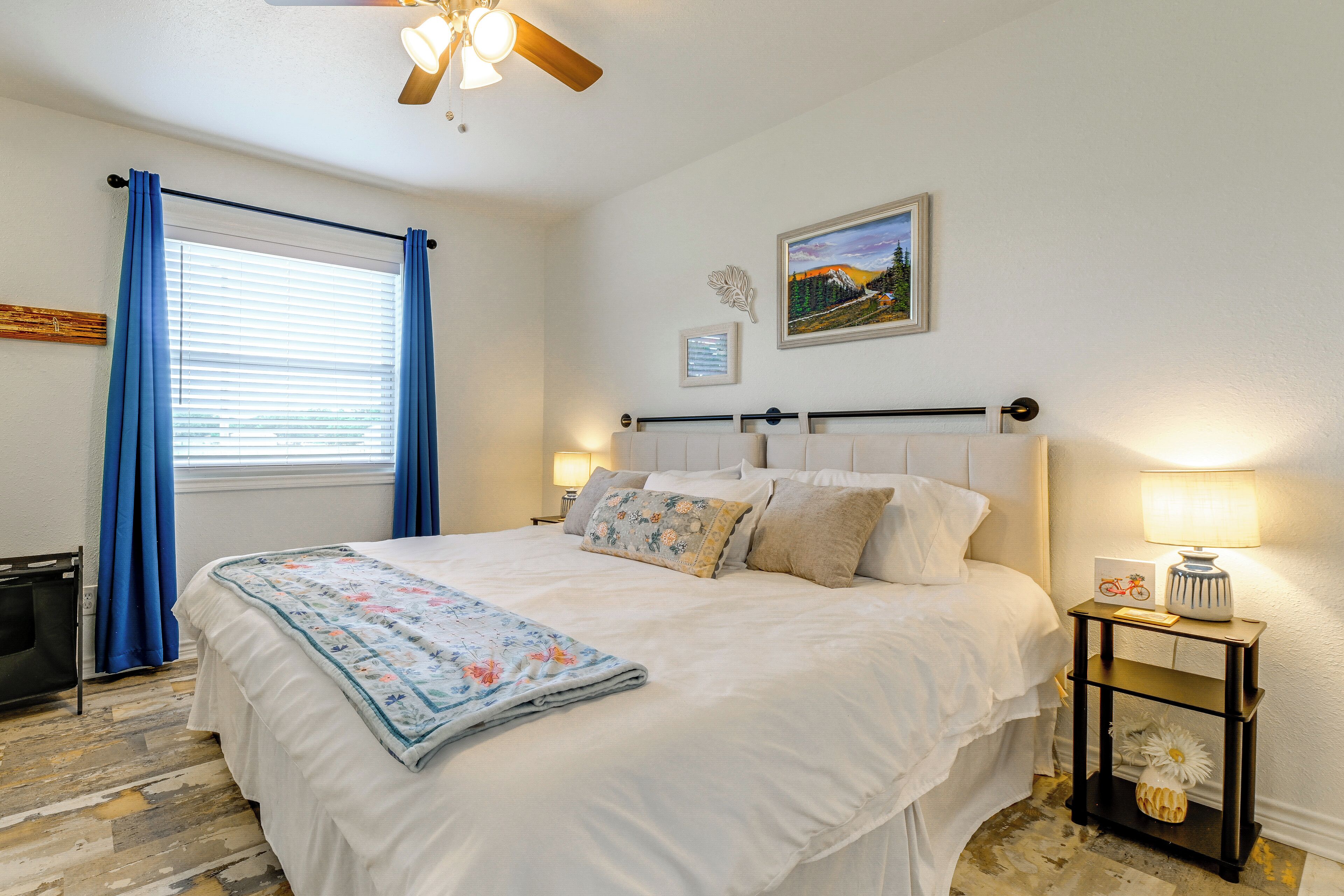 A bedroom with a bed, a nightstand, a ceiling fan, a window with curtains, and a wall-mounted picture.