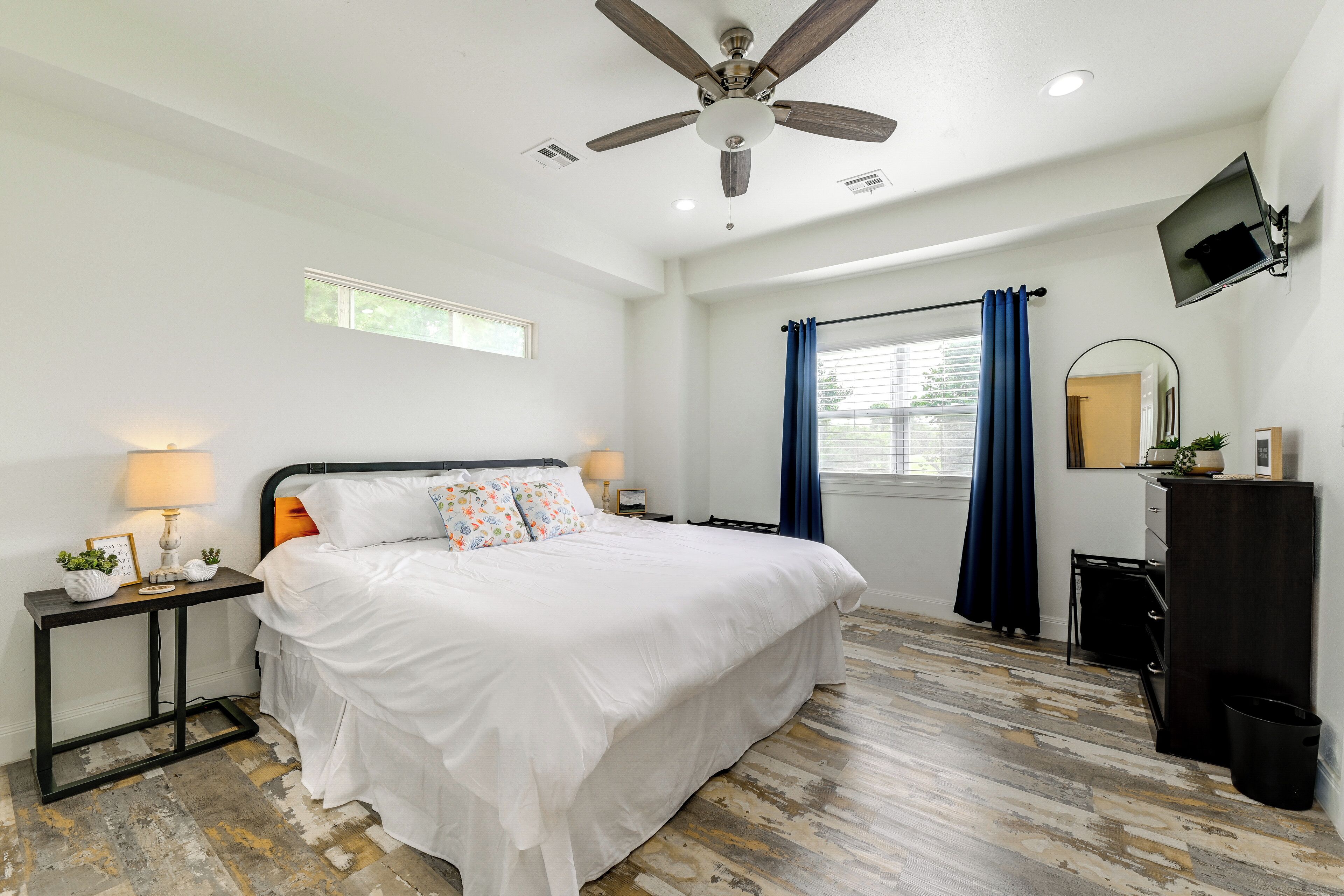 A bedroom with a bed, ceiling fan, TV, window, and wooden flooring.