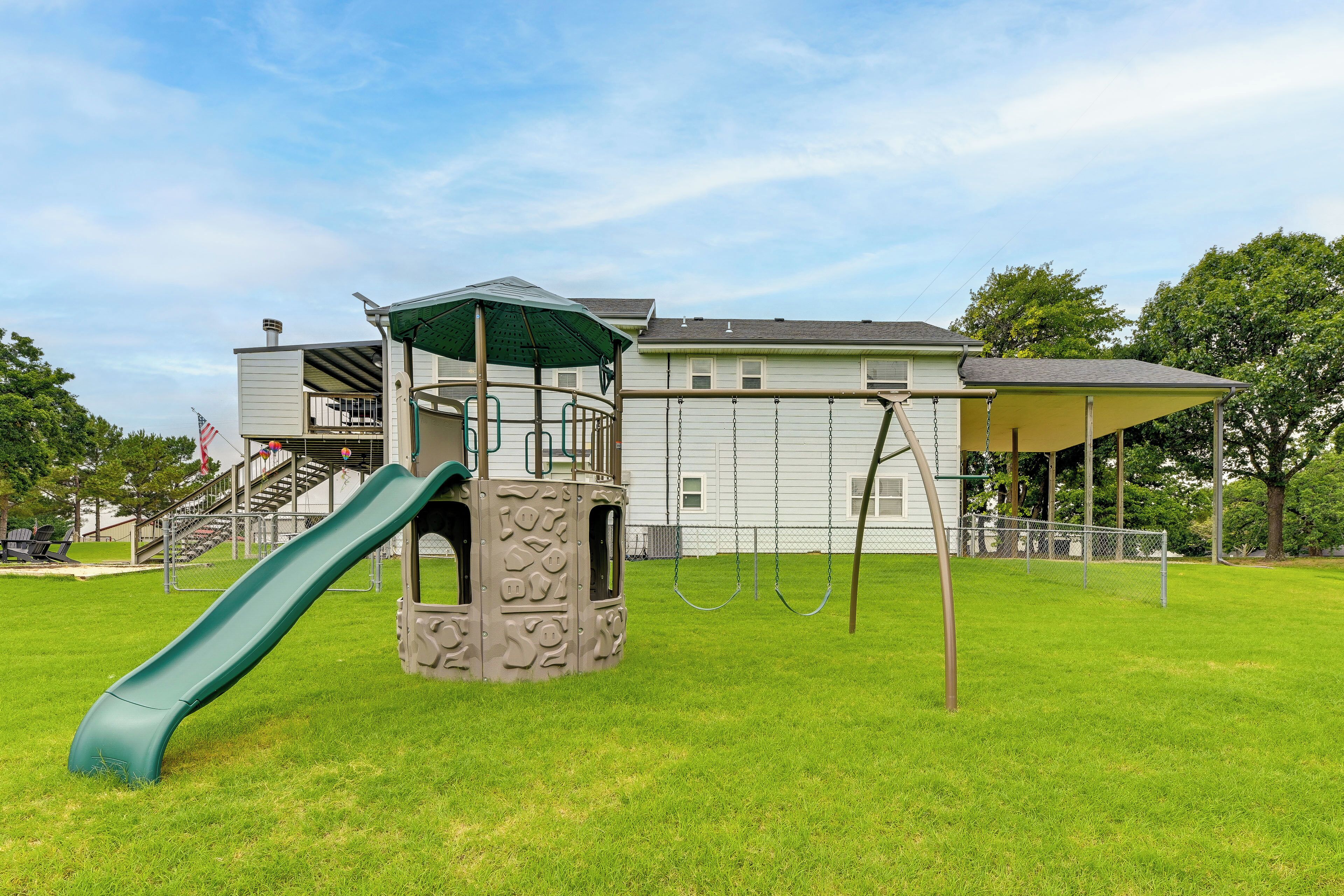 Un parque infantil con tobogán y columpios, rodeado de césped y un edificio con una estructura de juego techada.