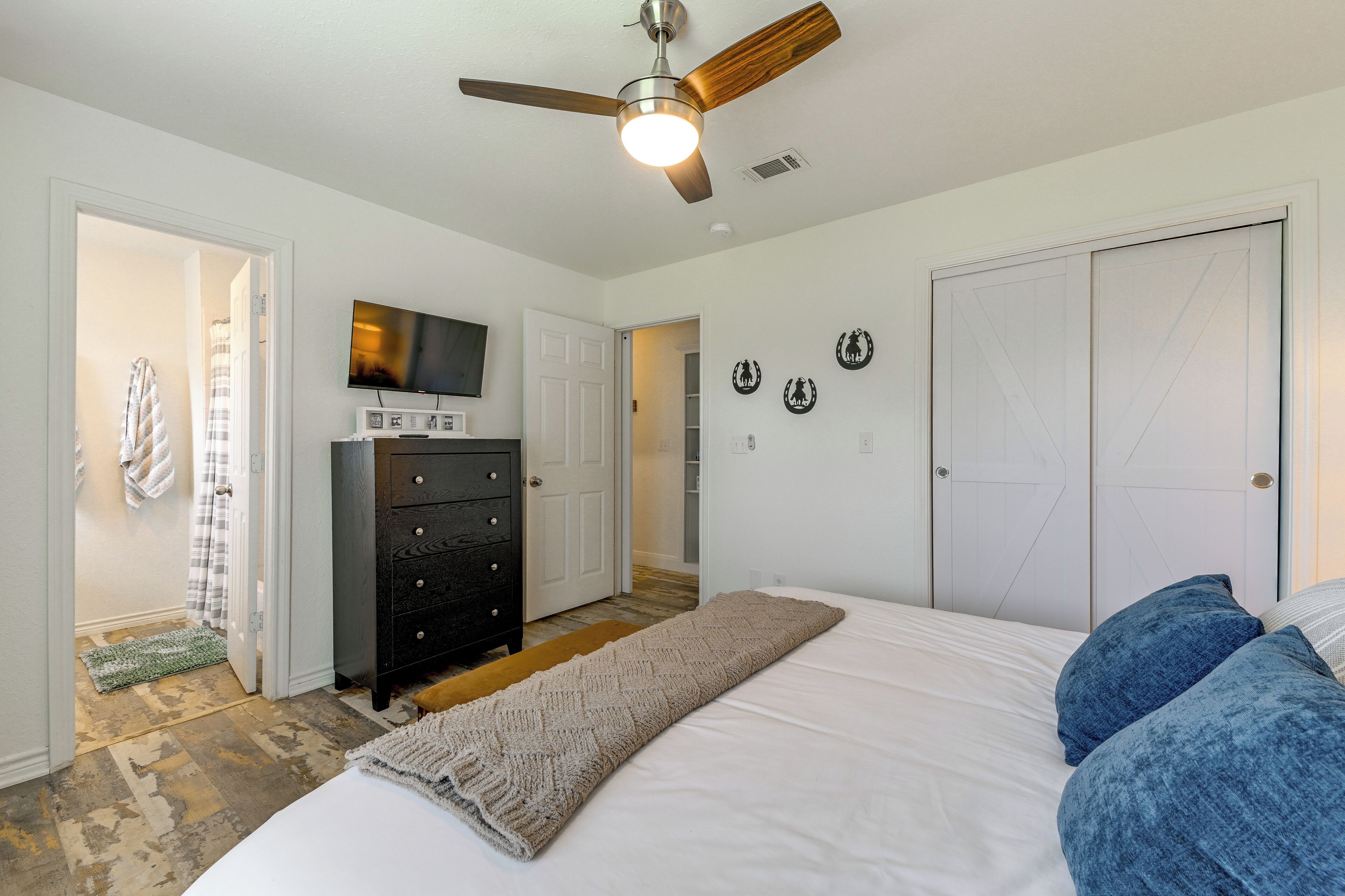 A bedroom with a bed, a ceiling fan, a dresser, a television, and a door leading to another room.