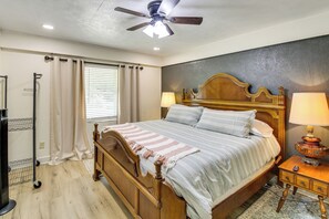 Ein Schlafzimmer mit einem hölzernen Bett, einem Deckenventilator, einem Nachttisch mit Lampe, einem Fenster mit Vorhängen und einem an der Wand montierten Fernseher.
