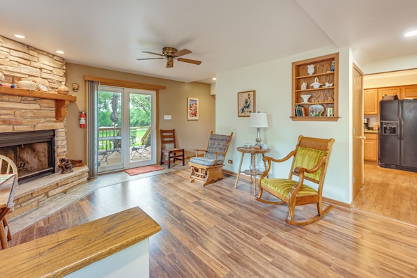 A spacious living area with a stone fireplace, wooden flooring, a ceiling fan, and a view through glass doors to an outdoor patio.