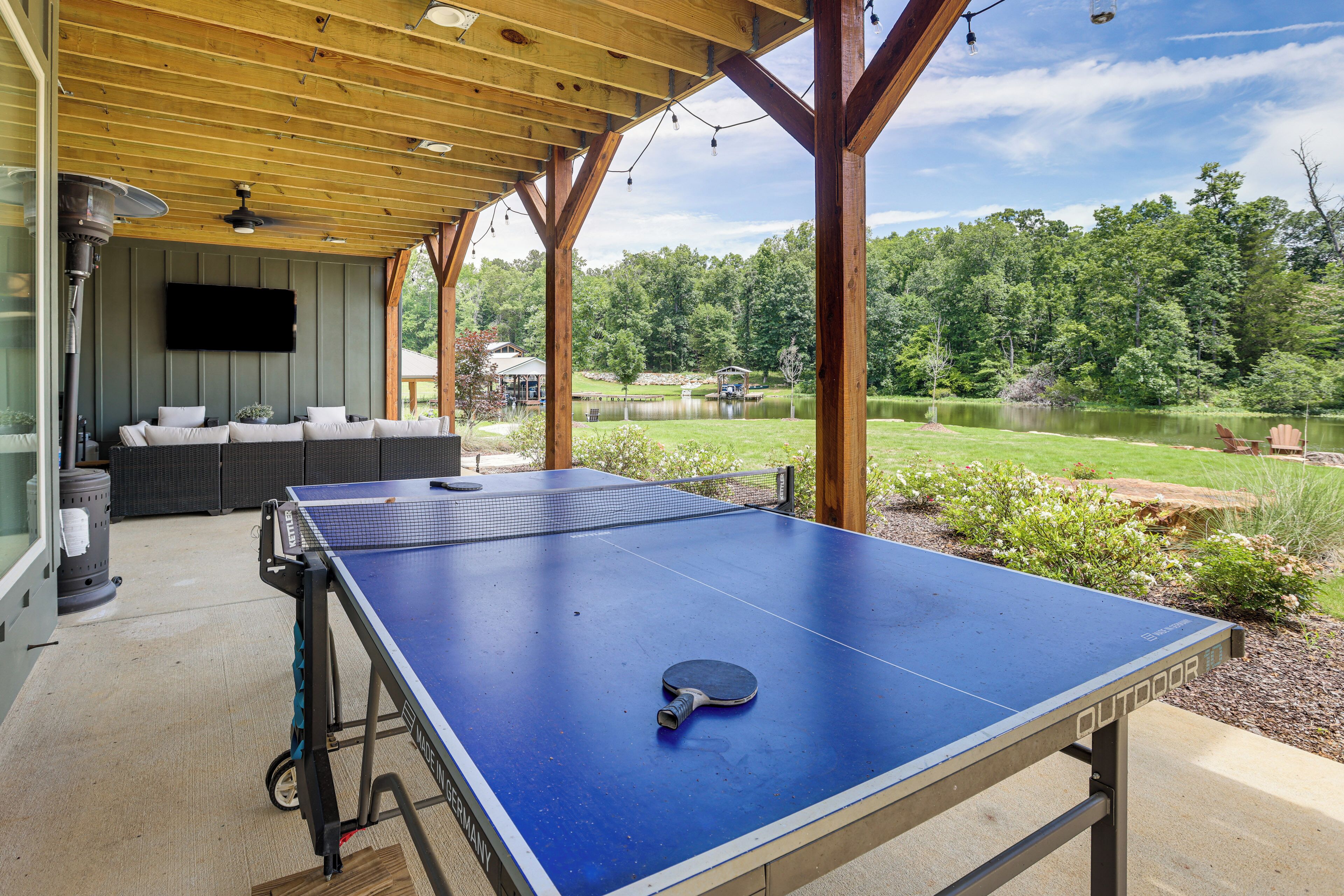 Una mesa de ping pong al aire libre con red, un televisor montado en una pared y una zona de estar cubierta con cojines.