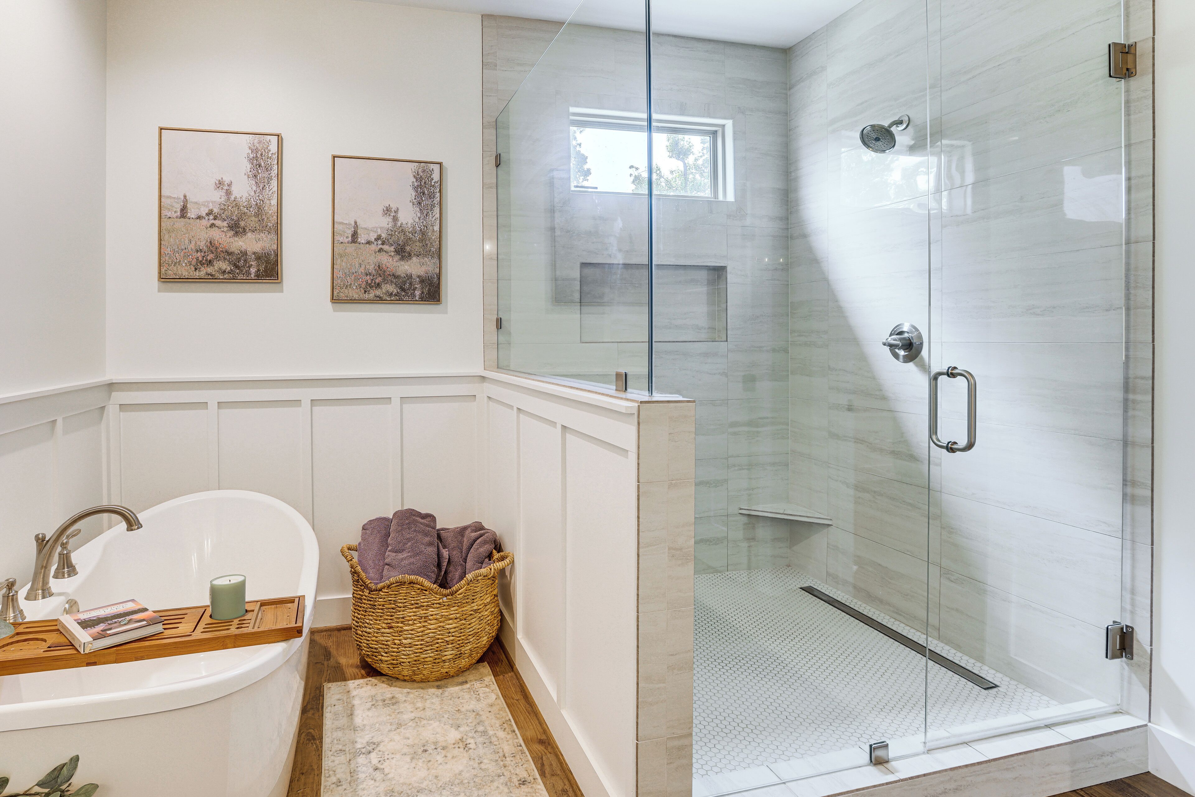 Un baño moderno con ducha de vidrio, una bañera independiente y dos cuadros con paisajes en la pared.