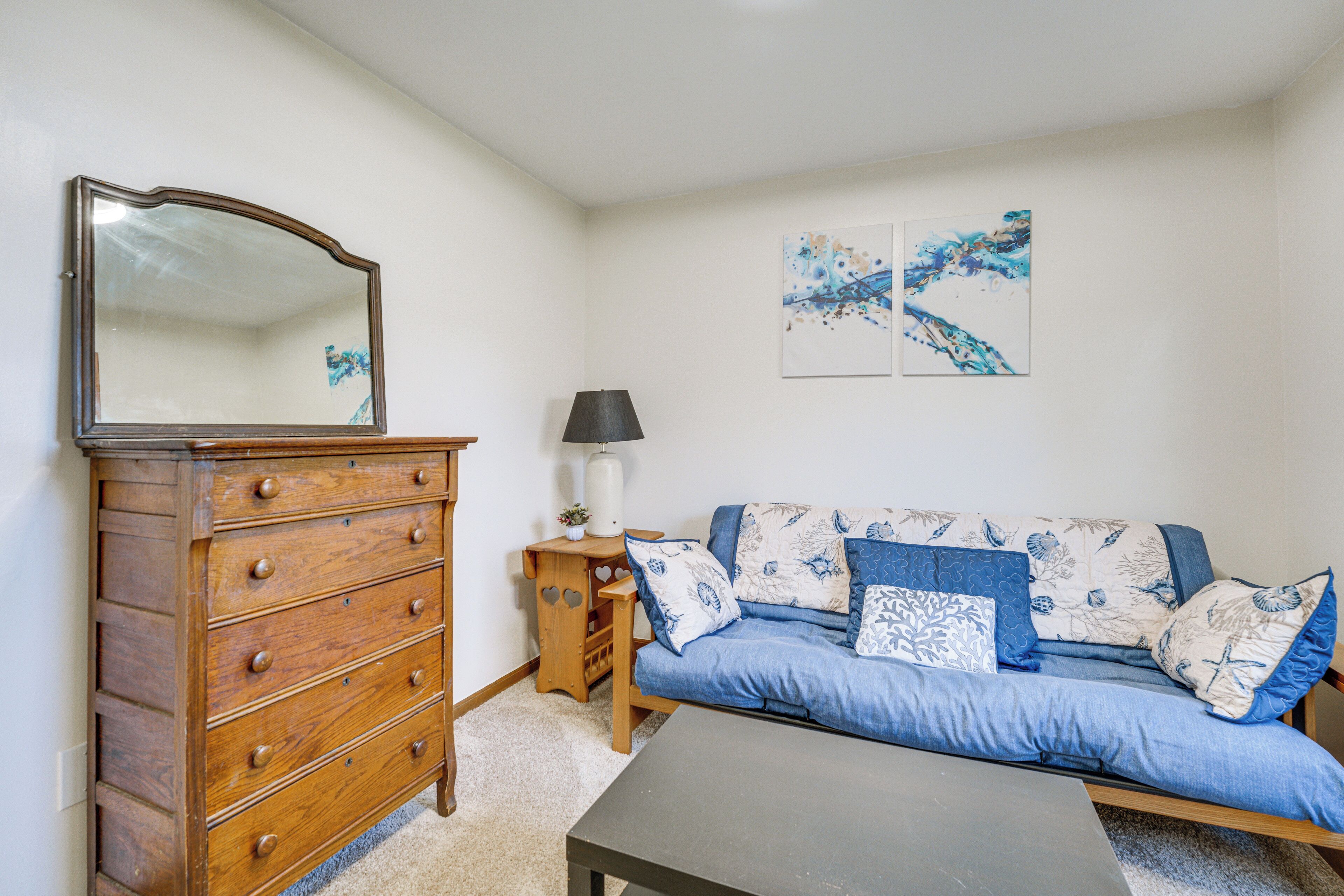 A room with a wooden dresser, a blue sofa with patterned cushions, a table, and wall art.