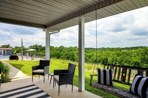 Een overdekt terras met stoelen en een schommel, met uitzicht op een gazon en bomen.