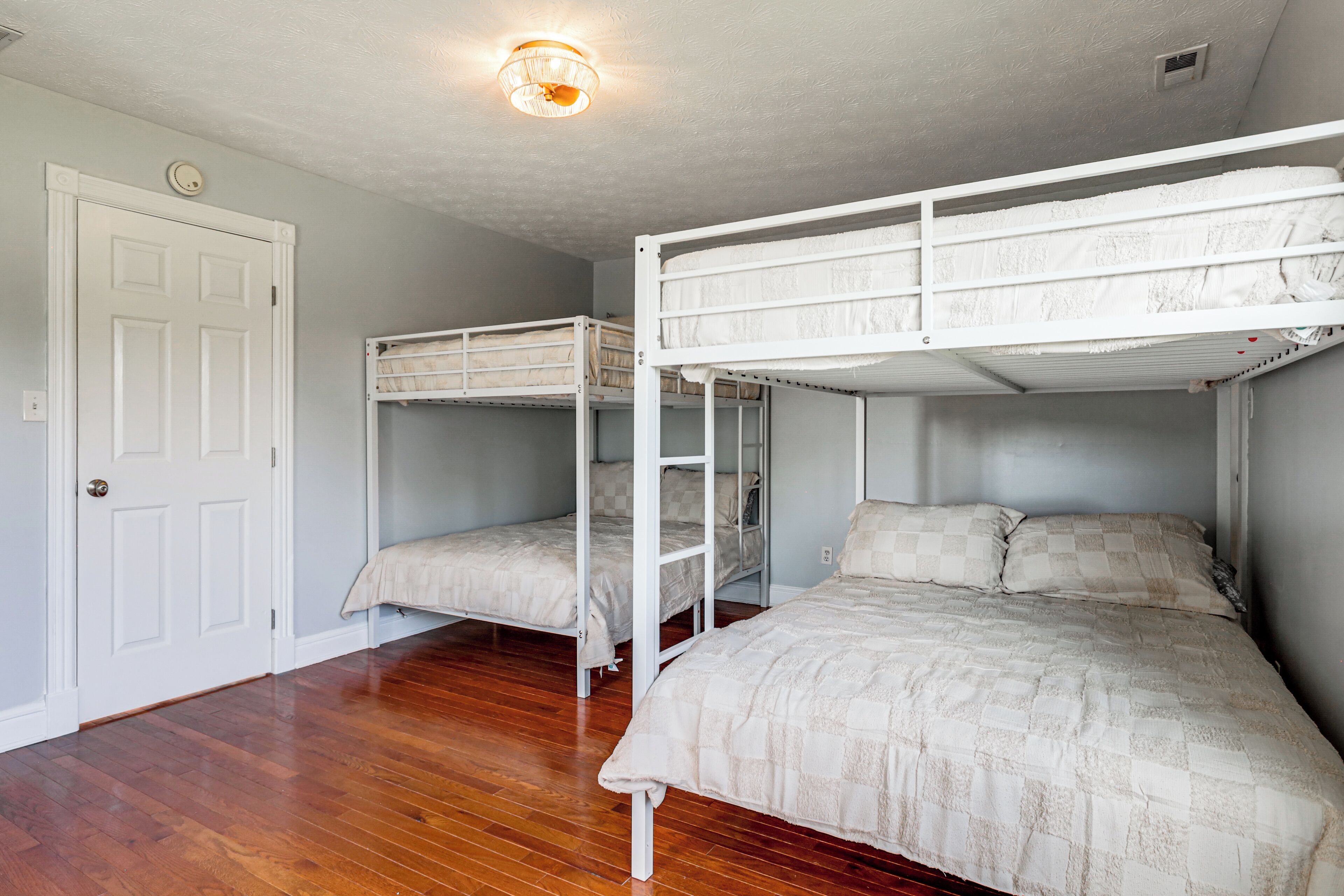 Une pièce avec deux lits superposés, un plancher en bois, une porte blanche et un luminaire au plafond.