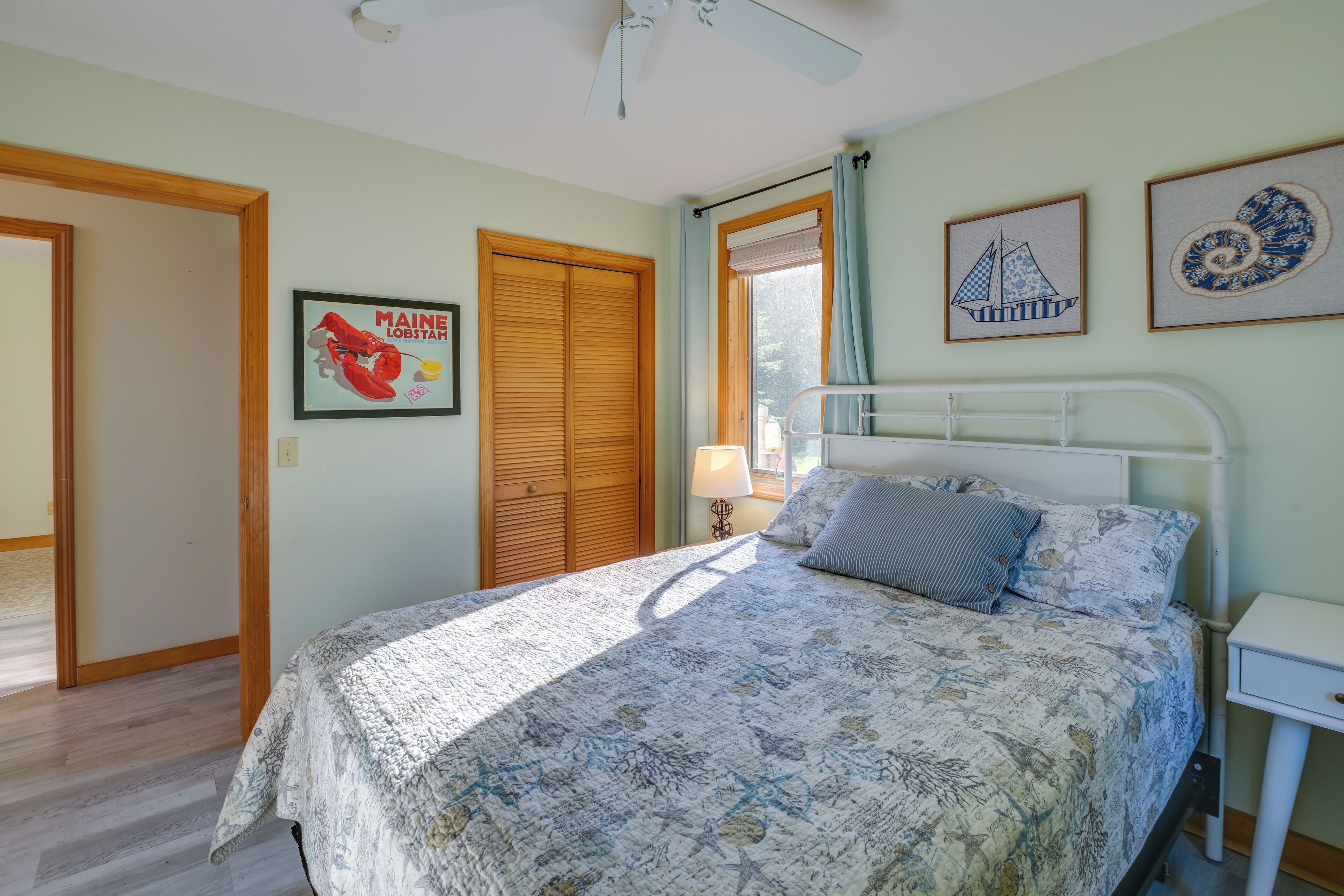 A bedroom with a bed, a nightstand, a window with curtains, a ceiling fan, and a door.