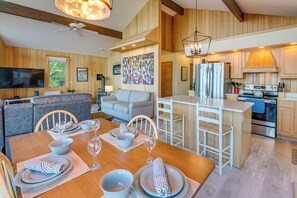 Una cocina moderna con armarios de madera, un comedor con una mesa de madera dispuesta para cuatro personas, y una sala de estar con sofá y televisor.