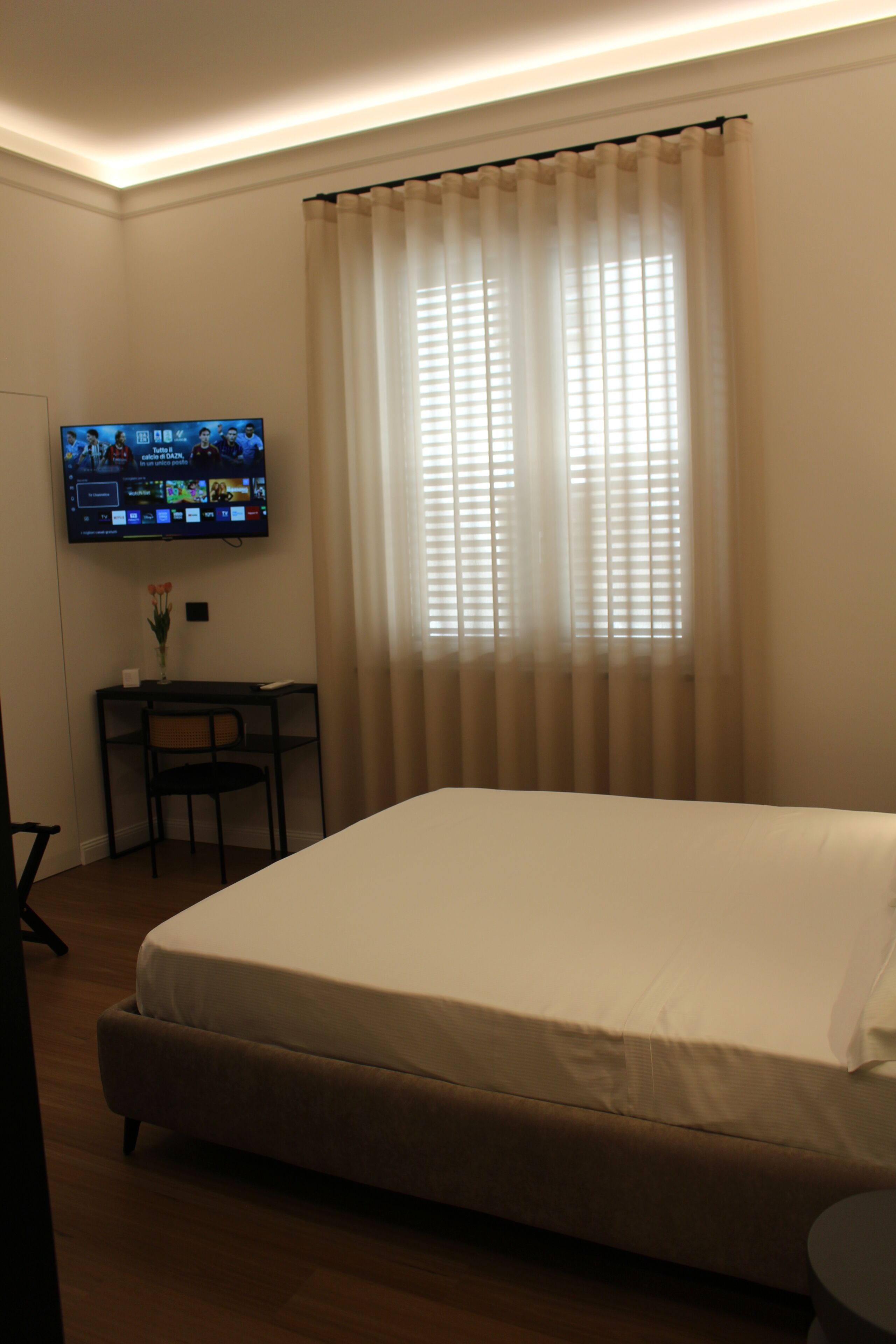 Camera d'albergo con un letto, una scrivania, una sedia, una televisione a muro e una finestra con tende.