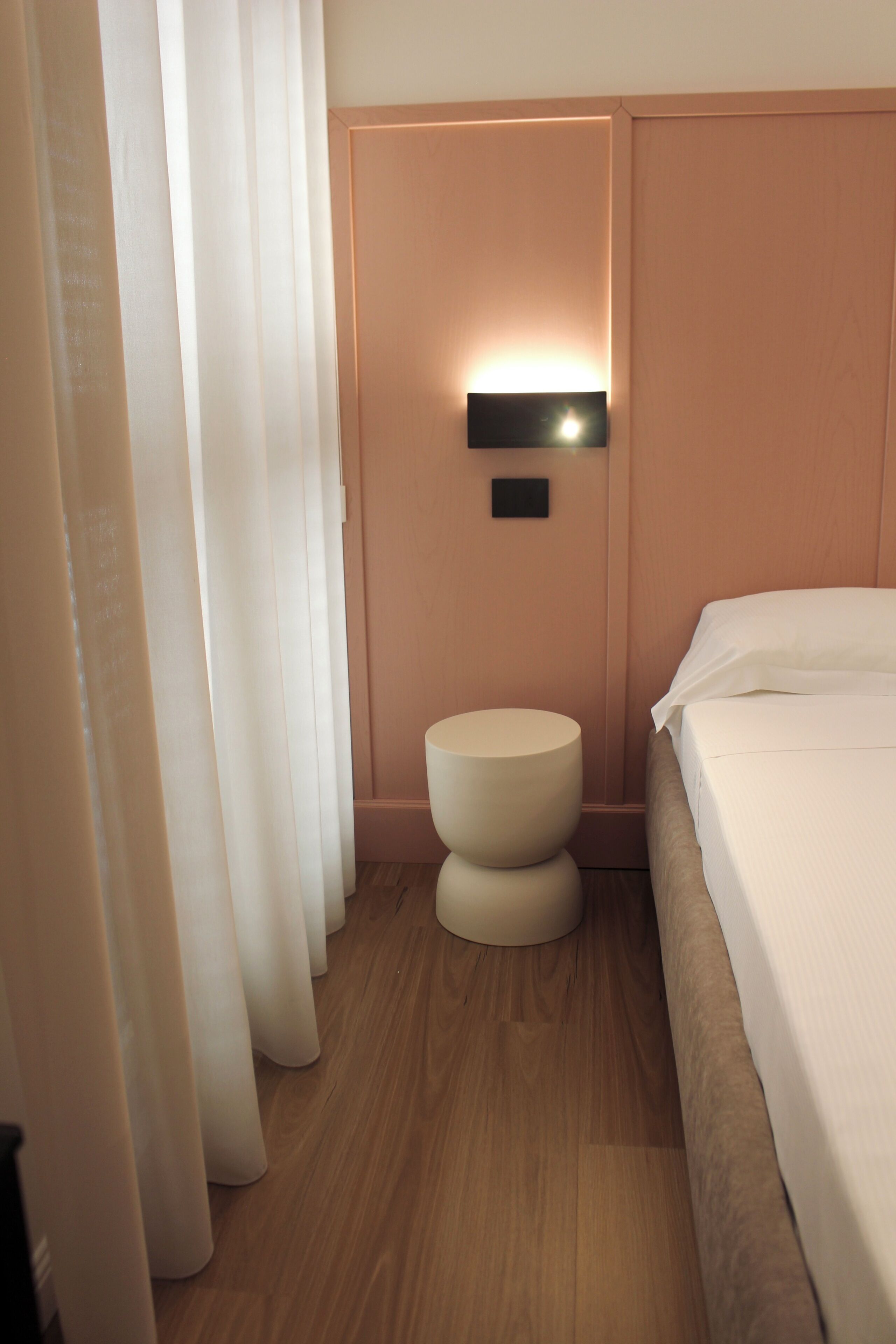 A hotel room with a bed, a bedside table, a wall-mounted light, and a curtain.