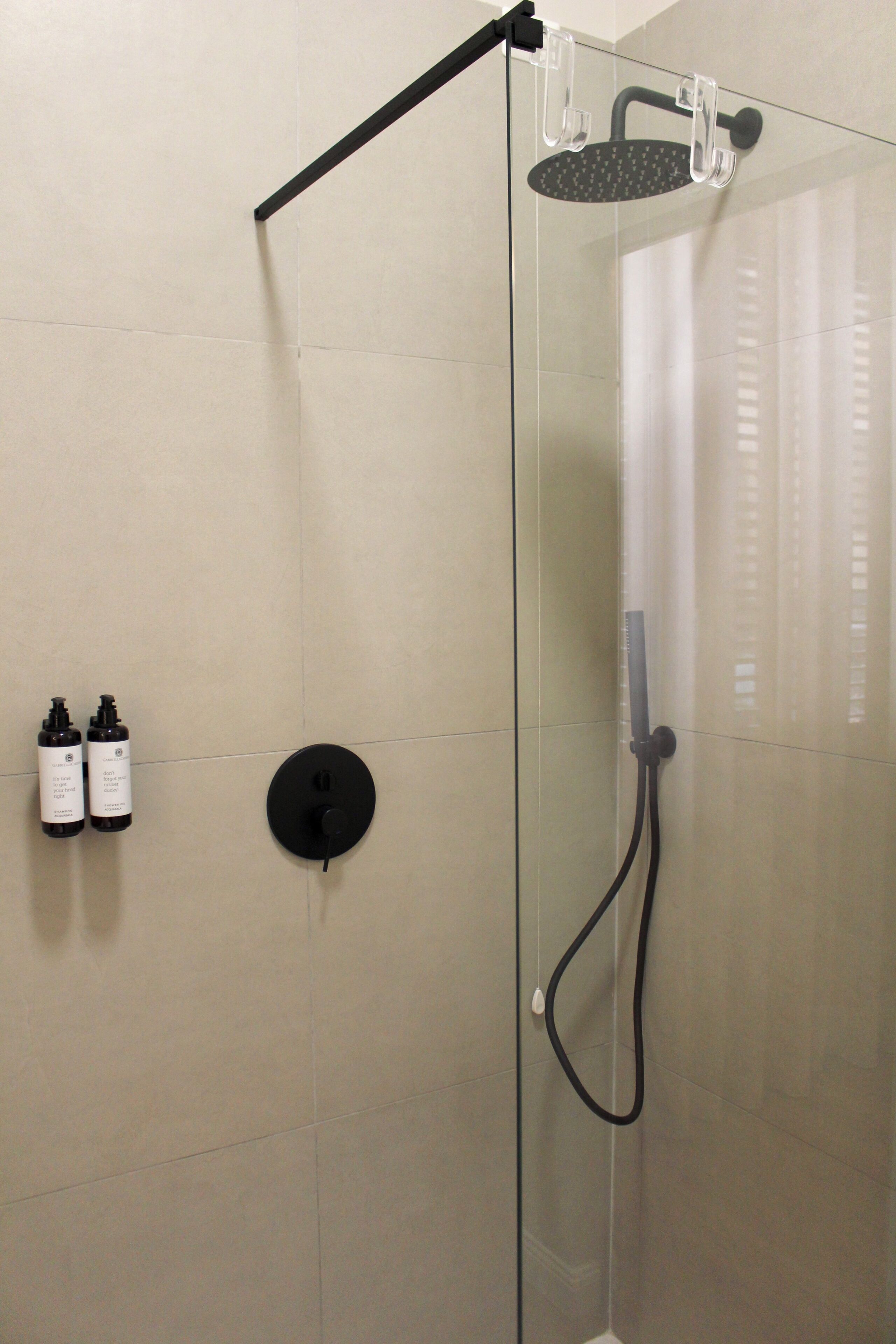 A glass-enclosed shower with a handheld showerhead, a handheld shower attachment, and two bottles of shampoo on the wall.
