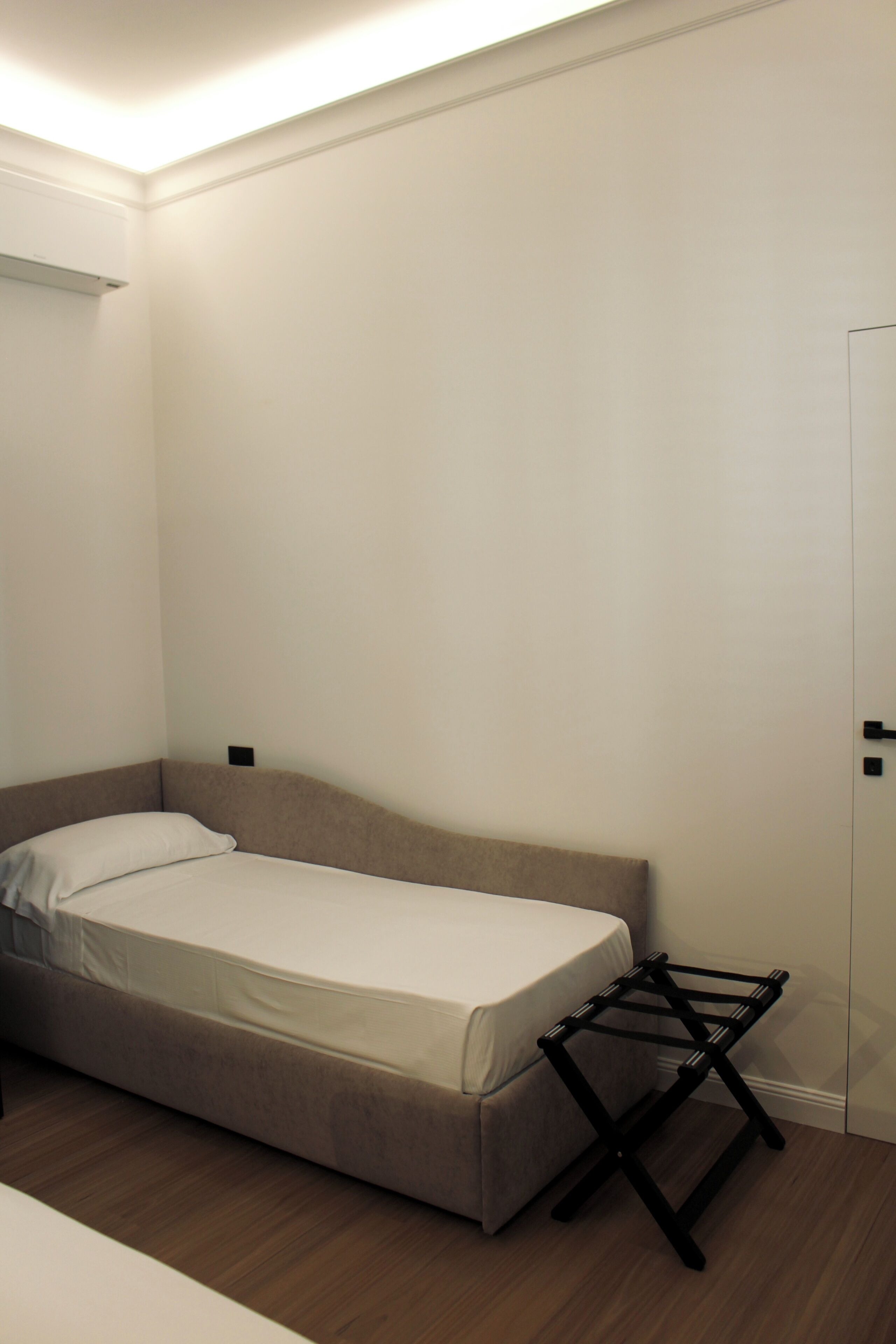 A small, single-bed room with a wooden floor, a bedside table, and a wall-mounted shelf.