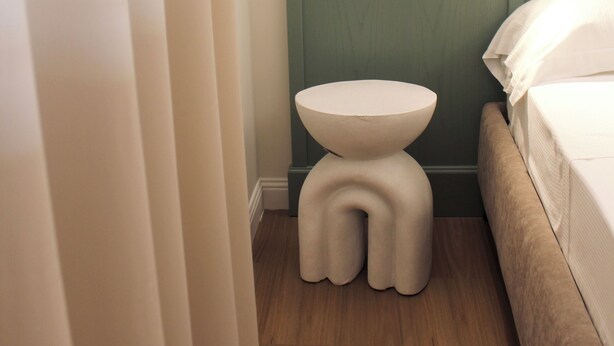 Un baño pequeño y moderno con piso de madera, un lavamanos blanco, una lámpara de pared y una cama con ropa de cama blanca.