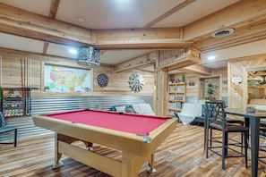 A pool table in a wooden room with a map on the wall, a dartboard, and shelves with books and games.