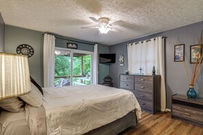 A bedroom with a bed, a ceiling fan, a dresser, a window with curtains, and a view of greenery outside.