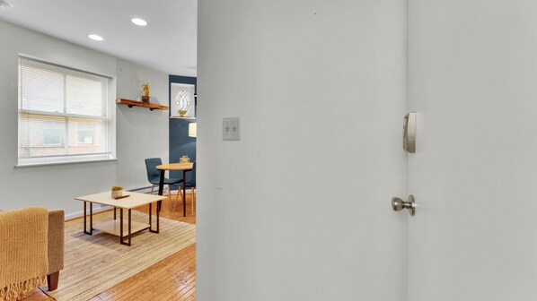 Quarto com piso de madeira, uma pequena mesa com cadeiras, uma janela com persianas e uma porta com puxador.