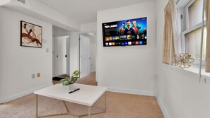 Uma sala de estar moderna com uma TV de tela plana na parede, uma mesa de centro branca com um controle remoto, uma planta em vaso e uma obra de arte emoldurada na parede.