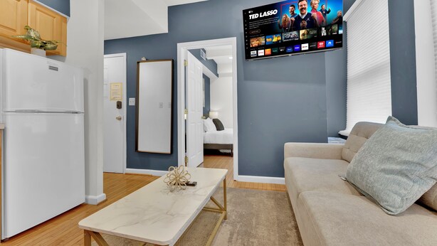 A modern living room with a flat-screen TV mounted on the wall, a sofa, a coffee table, and a kitchen area with a refrigerator. - Apartmint at 242 S 17th St Philadelphia (Philadelphia)