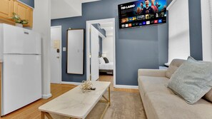 Una sala de estar moderna con un televisor de pantalla plana montado en la pared, un sofá, una mesa de centro y una zona de cocina con un refrigerador.