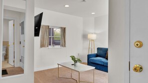 Una habitación con una puerta blanca, un sofá azul, una mesa de centro blanca, una lámpara de pie y un televisor montado en la pared.