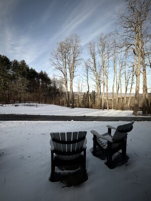 Zwei Adirondack-Stühle auf verschneitem Grund, mit Blick auf einen Wald und eine Straße im Hintergrund.