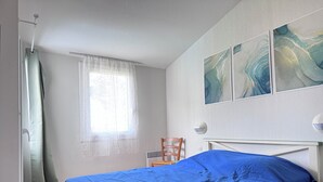Ett sovrum med en säng, en stol, ett fönster med tunna gardiner och abstrakt konst på väggen.