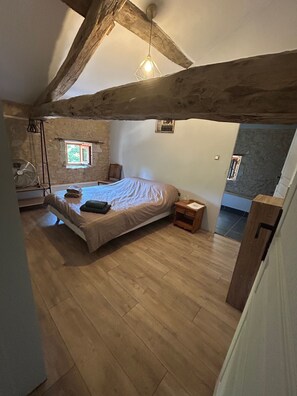 Ein Schlafzimmer mit einem Bett, Holzbalken, einem Fenster, einem Ventilator und einem kleinen Tisch.
