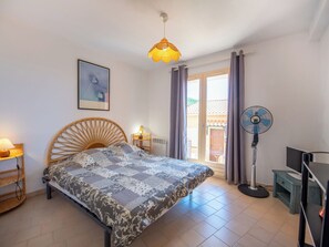 A bedroom with a bed, a standing fan, a desk with a computer, a lamp, and a window with curtains. - Les Sauvagières II by Interhome (Cavalaire)