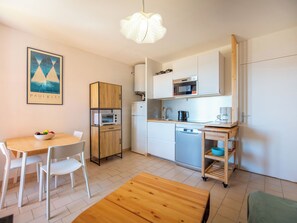 A compact kitchen with a microwave, oven, and coffee maker, a dining area with a wooden table and chairs, and a framed picture on the wall. - Les Sauvagières II by Interhome (Cavalaire)