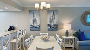 Uma sala de jantar com uma mesa, cadeiras, uma cesta de frutas e duas pinturas de barcos à vela.