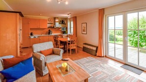 Amplia sala de estar con sofá, una mesa de centro y una mesa de comedor. Se ve una cocina al fondo con armarios y un microondas. La habitación tiene ventanas grandes con vistas a zonas verdes.