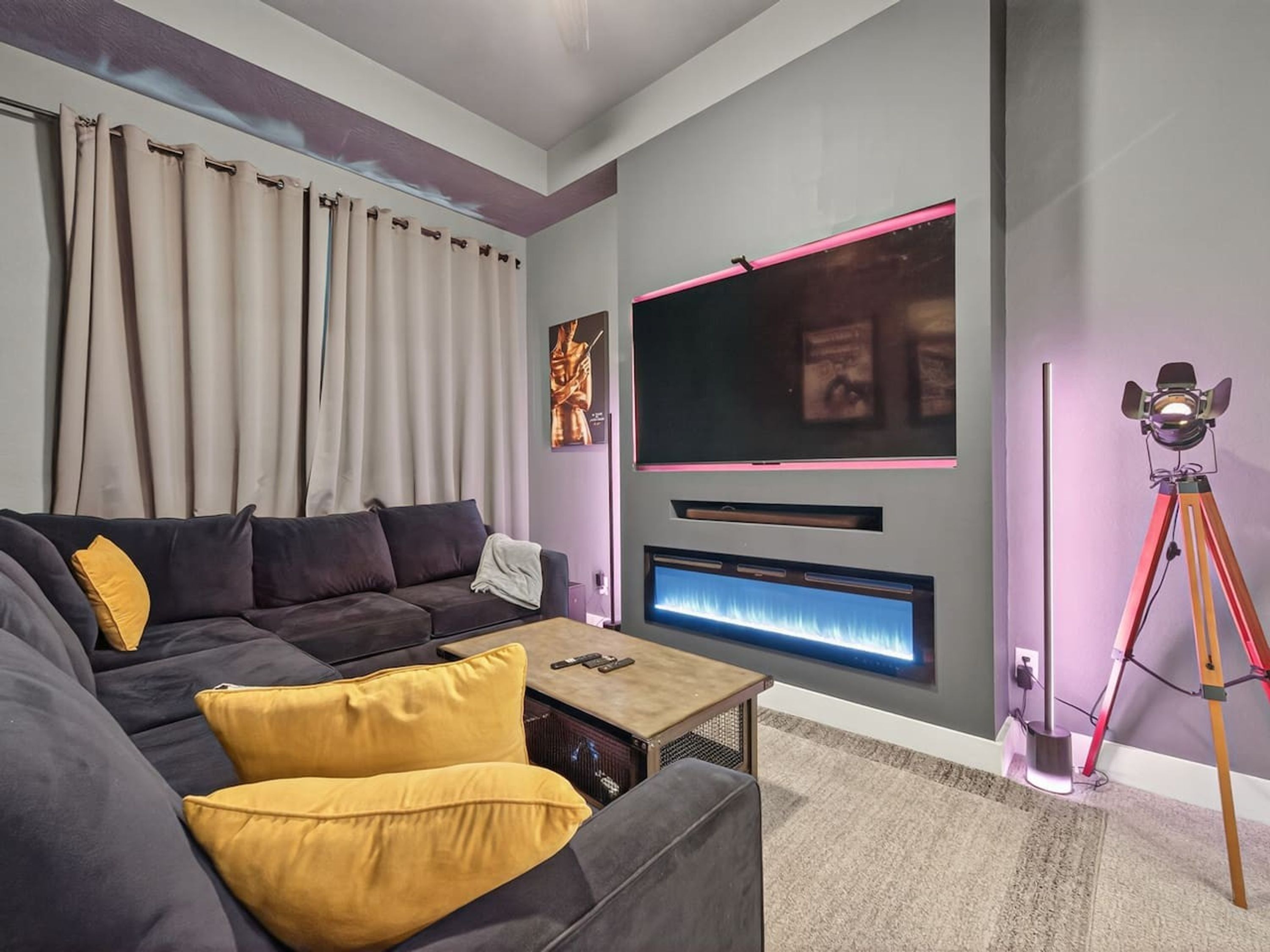 Una sala de estar moderna con un sofá gris oscuro, cojines amarillos, una mesa de centro, un televisor de pantalla plana, un sistema de sonido integrado en la pared y una lámpara con trípode.
