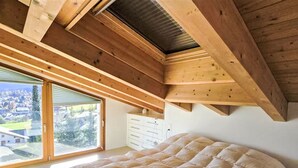 Quarto com cama grande, vigas de madeira no teto, porta de vidro deslizante e vista para uma cidade.
