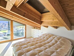 A bedroom with a large bed, wooden ceiling beams, a sliding glass door, and a view of a town. - Casa Jörger by Interhome (Flims)