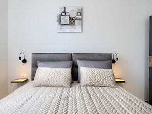 Ein Bett mit weißer Steppdecke, zwei grauen Kissen, einem grauen Kopfteil und zwei Nachttischlampen.