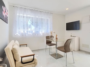 Ein modernes Wohnzimmer mit einer Couch, einem Glastisch, Stühlen, einem Fernseher und einem Fenster mit transparenten Vorhängen.
