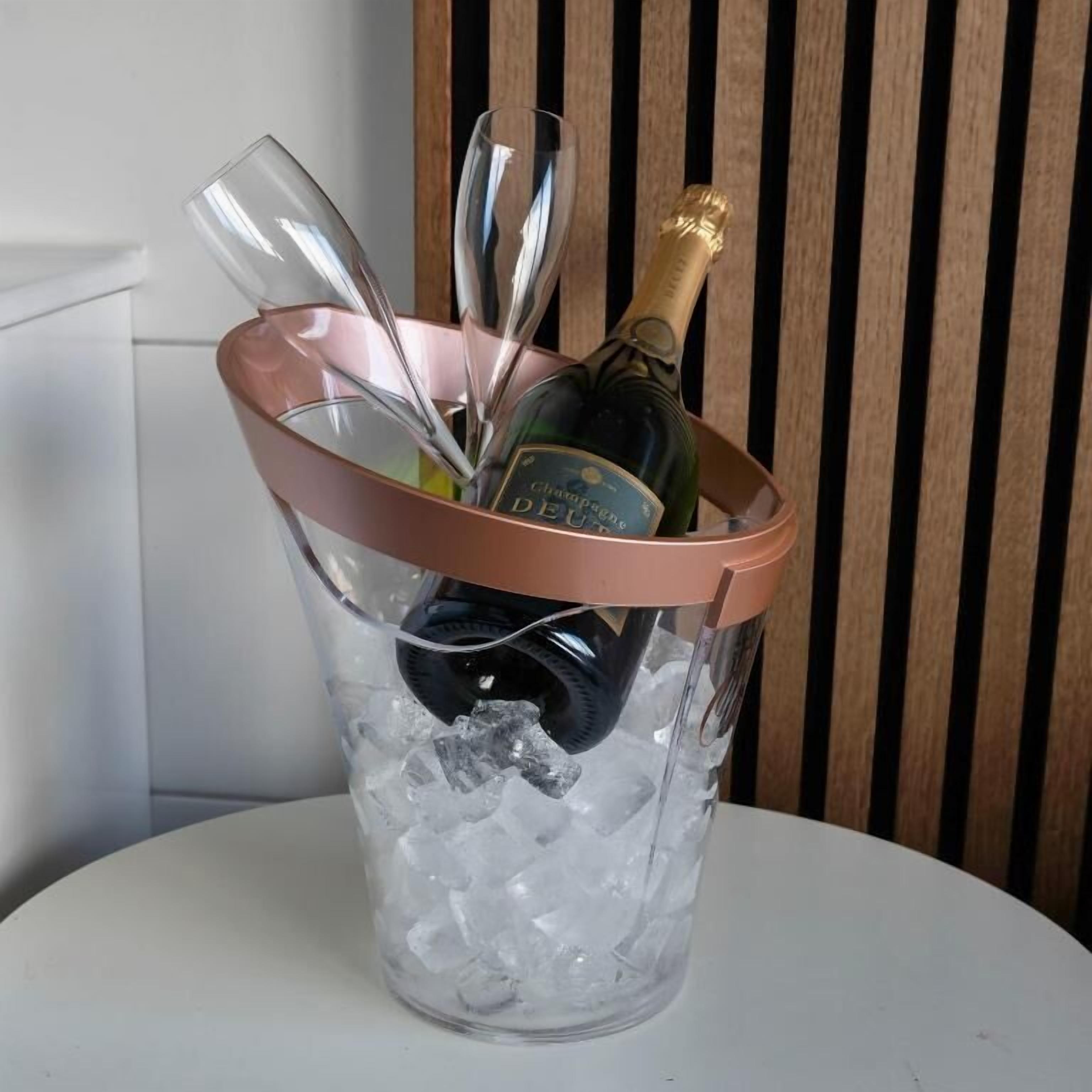 Um balde de vidro transparente com alça de cortiça contendo uma garrafa de champanhe e vários copos de vinho, sobre uma mesa com gelo.