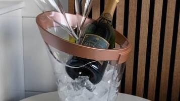 Um balde de vidro transparente com alça de cortiça contendo uma garrafa de champanhe e vários copos de vinho, sobre uma mesa com gelo.